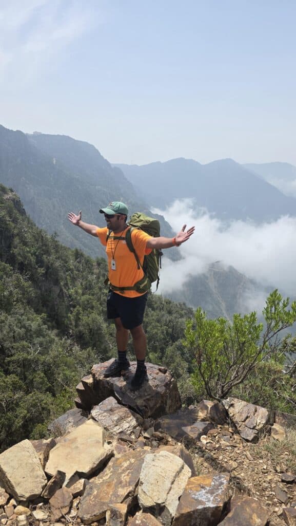 Climber standing on rocky mountain peak with arms outstretched, overlooking lush green valleys and m.