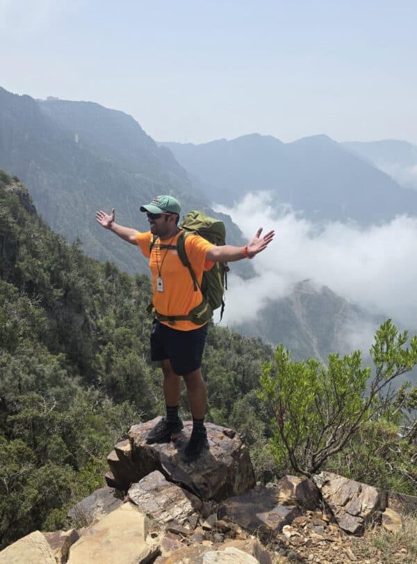 Climber standing on rocky mountain peak with arms outstretched, overlooking lush green valleys and m.